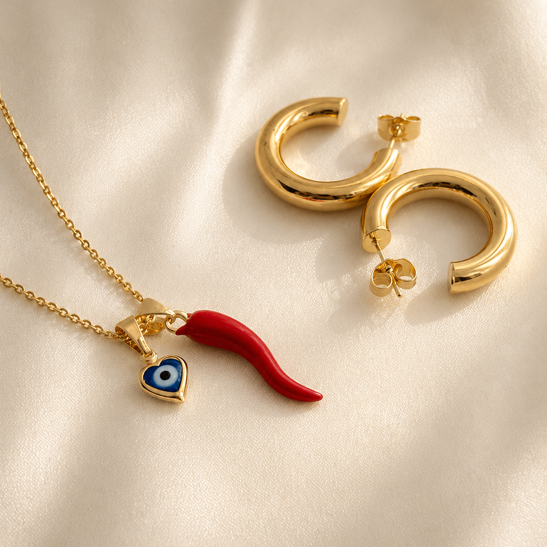 Gold filled necklace with red italian horn and blue evil eye pendant and gold hoop earrings on a beige fabric background