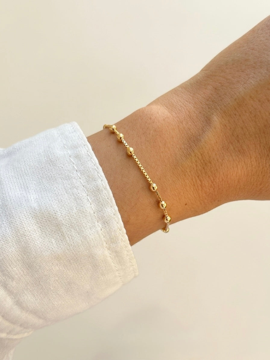 A delicate gold beaded chain bracelet worn on a person's wrist, paired with a white linen sleeve.