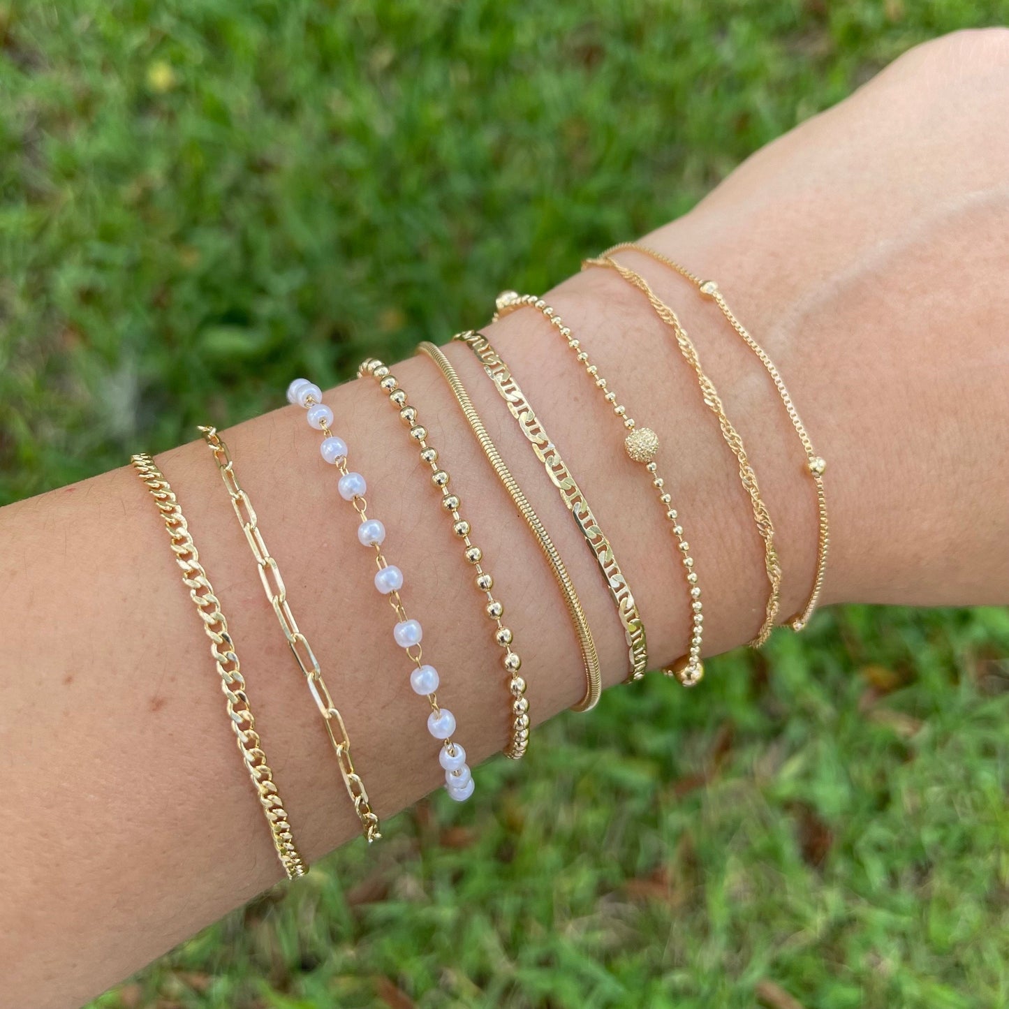 Dainty bracelets, satellite chain, singapur chain, beaded satellite bracelet, mariner bracelet, snake bracelet, beaded bracelet, pearl bracelet, paperclip bracelet, curb chain bracelet all in 18k gold filled on wrist and grass background.