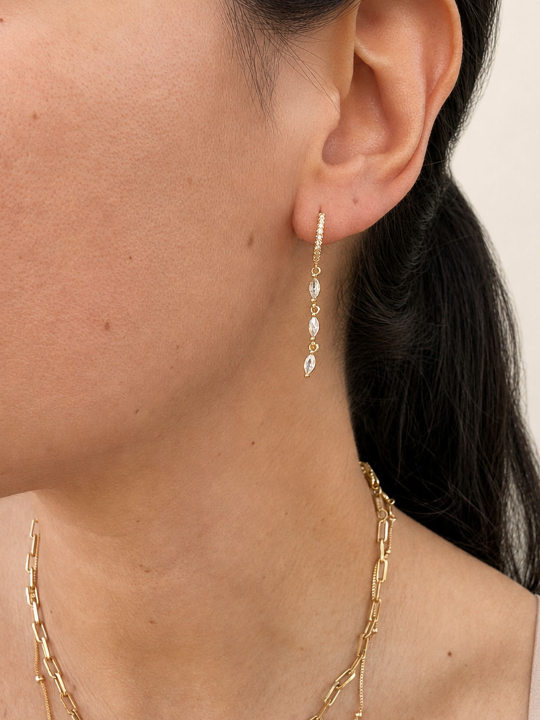 A close-up of a model's ear wearing a delicate gold huggie hoop earring with three small dangling clear teardrop-shaped stones. The model also wears layered gold necklaces against a soft beige background.