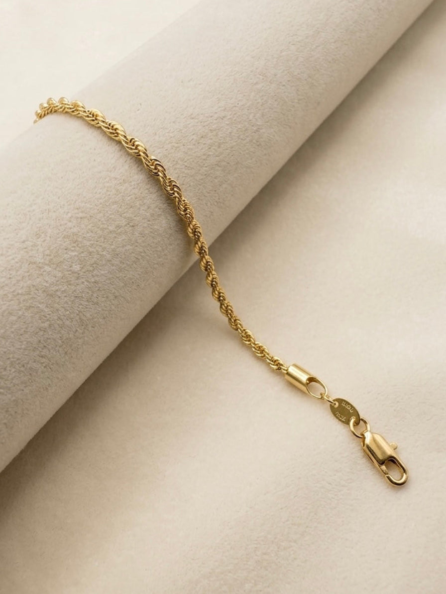 A delicate gold rope chain bracelet with a lobster clasp, displayed on a cream-colored textured fabric background.