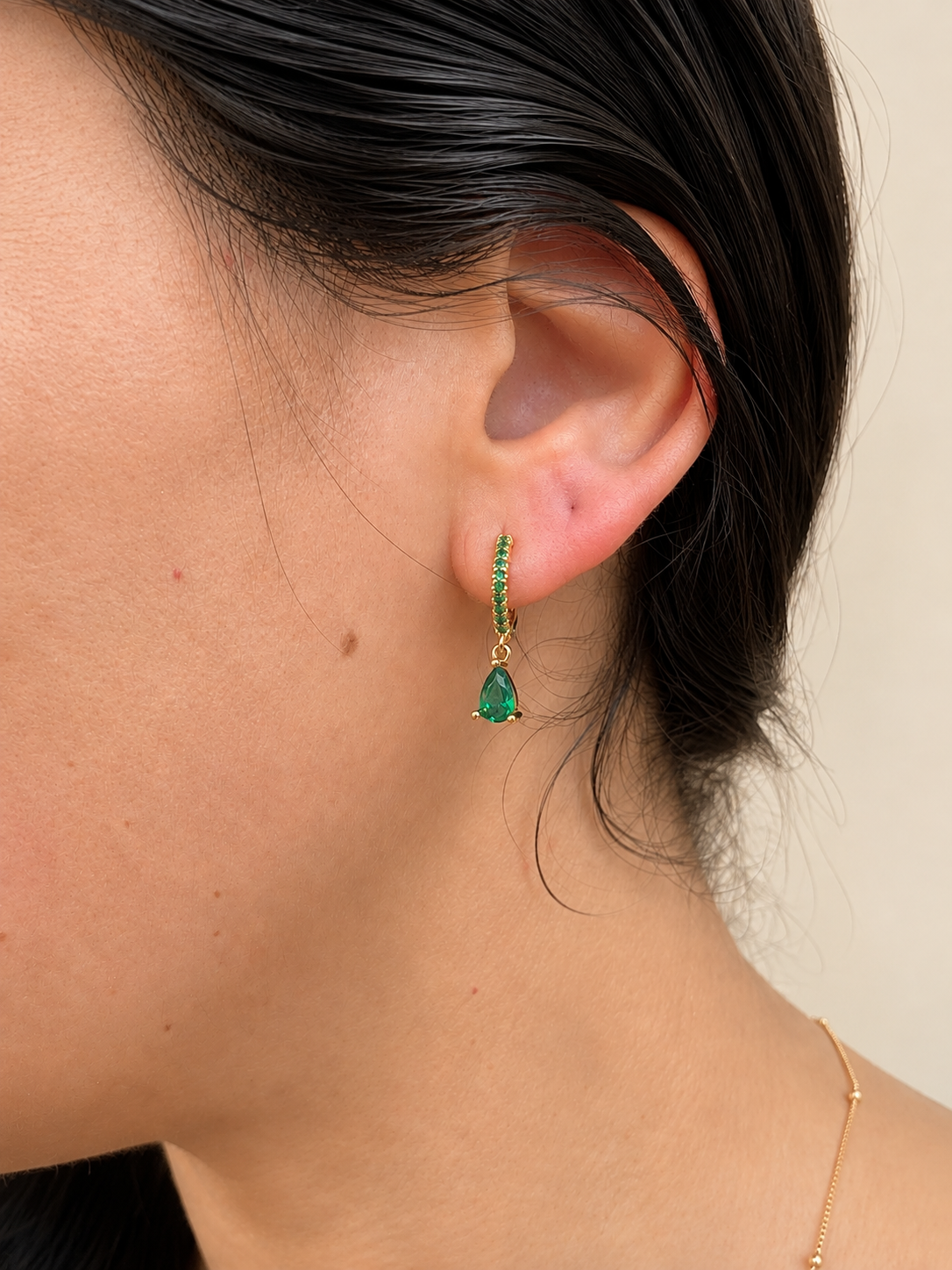 A close-up of a model's ear wearing a delicate gold huggie hoop earring adorned with small green stones and a teardrop-shaped green gemstone dangle.
