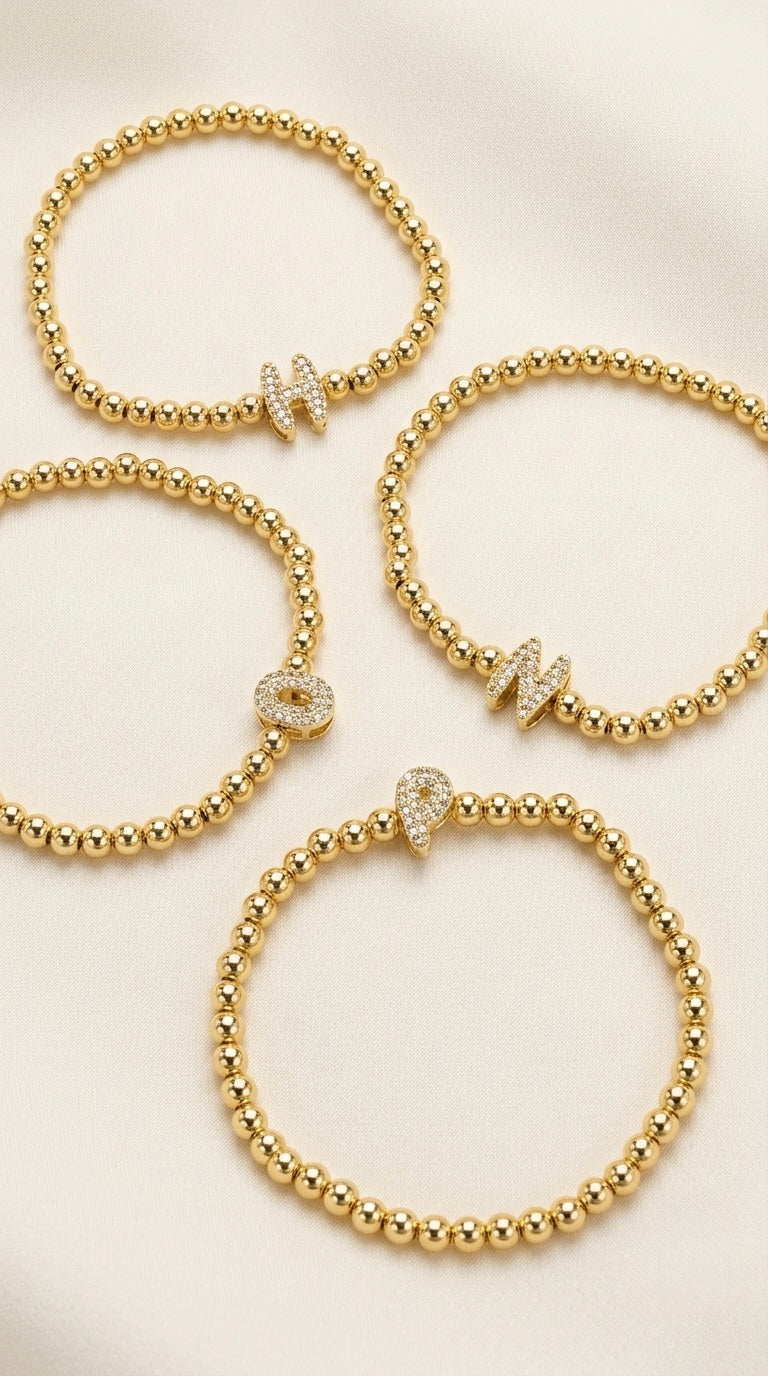 Four gold plated beaded stretch bracelets arranged on a light cream background, each featuring a small letter charm set with clear stones.