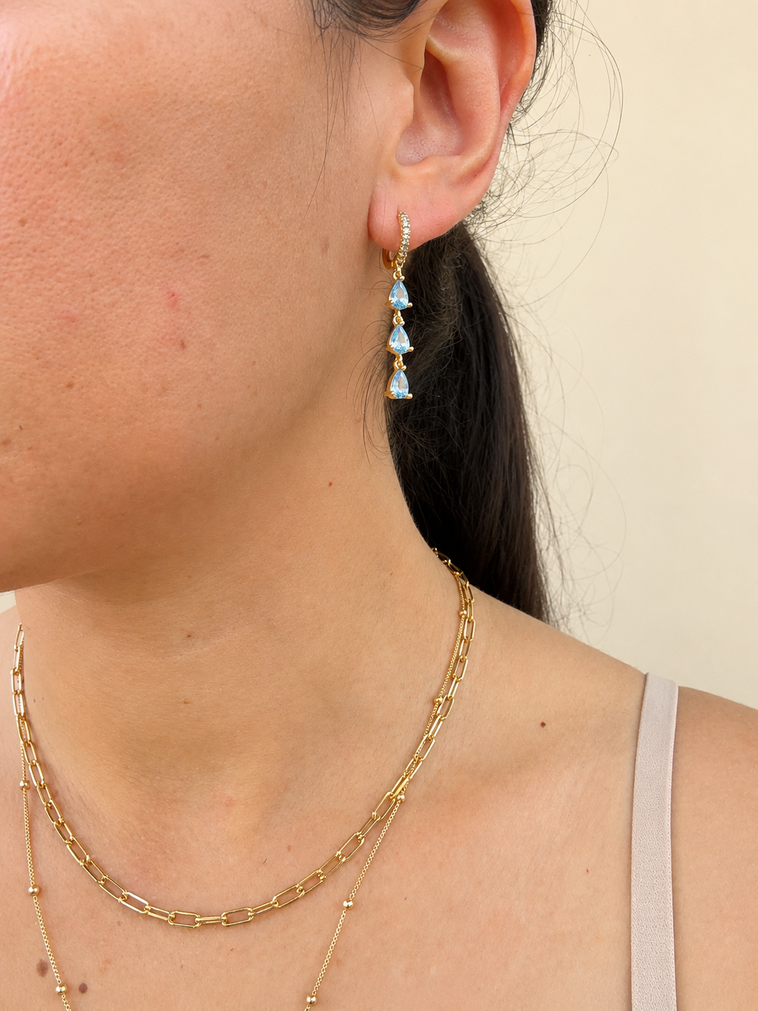A close-up of a woman's ear wearing a gold drop earring with three light blue teardrop-shaped stones, along with layered gold necklaces and a beige tank top against a plain beige background.
