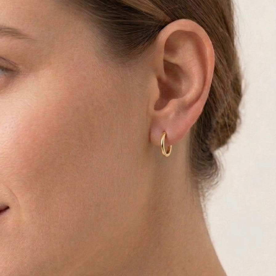 A close-up side profile of a woman wearing a small gold huggie hoop earring on her earlobe, against a clean off-white background.