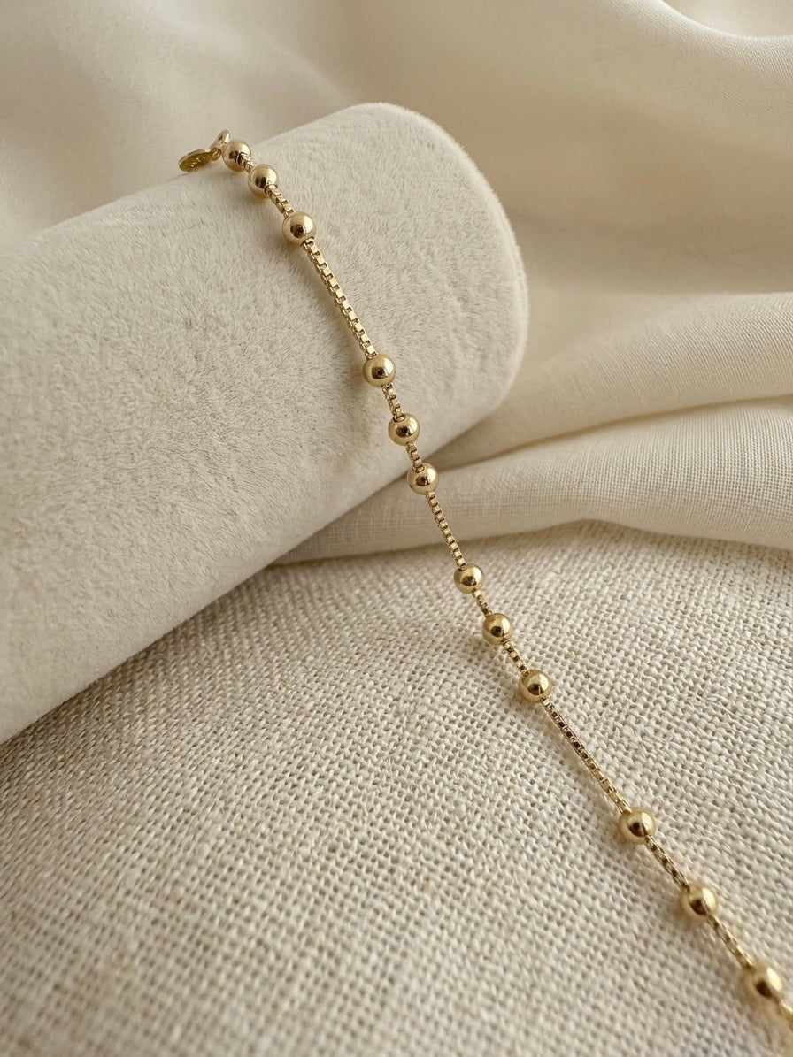 A delicate gold filled beaded chain bracelet draped over a cream-colored fabric roll on a textured linen surface.