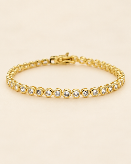 Gold-tone bezel tennis bracelet with round clear stones on a soft cream background.