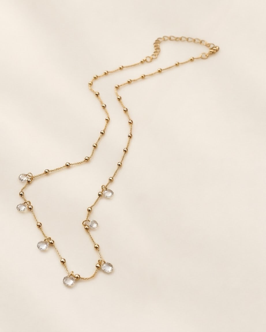 A delicate gold chain necklace with small bead accents and clear faceted crystal drops, displayed on a light cream background.