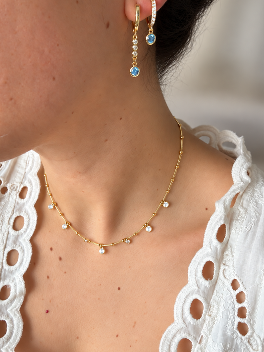 Close-up of a model wearing a delicate gold necklace with small clear stones and a matching gold drop earring with a light blue gemstone.
