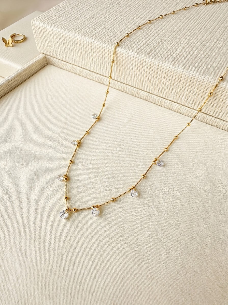 A delicate gold chain necklace with small clear crystal drops, displayed on a cream-colored textured jewelry box.