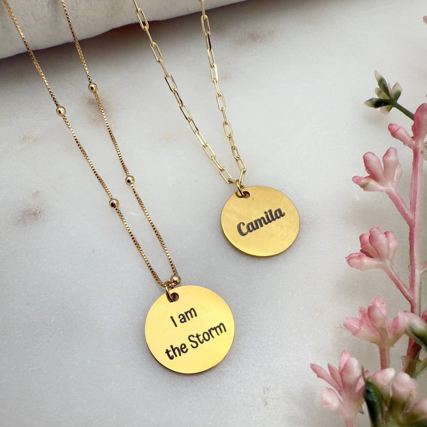 Two gold necklaces with engraved names on a light background with pink flowers.