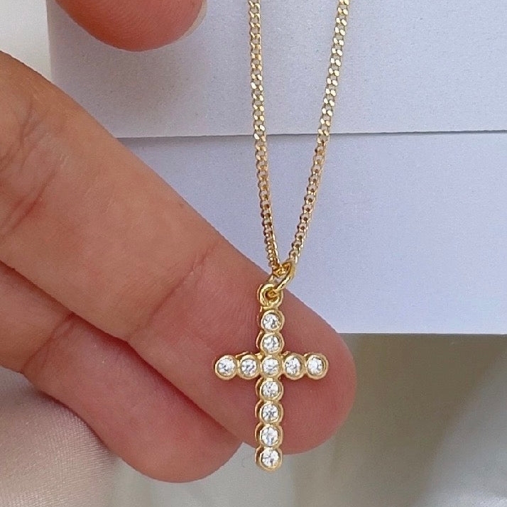 Gold cross necklace with clear stones held between fingers against a light background
