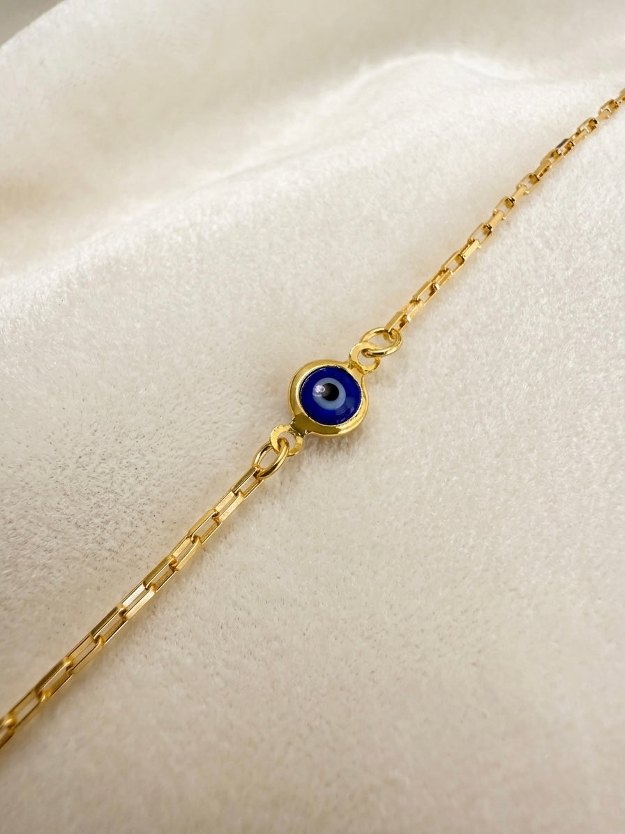 A gold filled paperclip chain bracelet featuring a small circular blue evil eye displayed on a soft, textured cream fabric background.