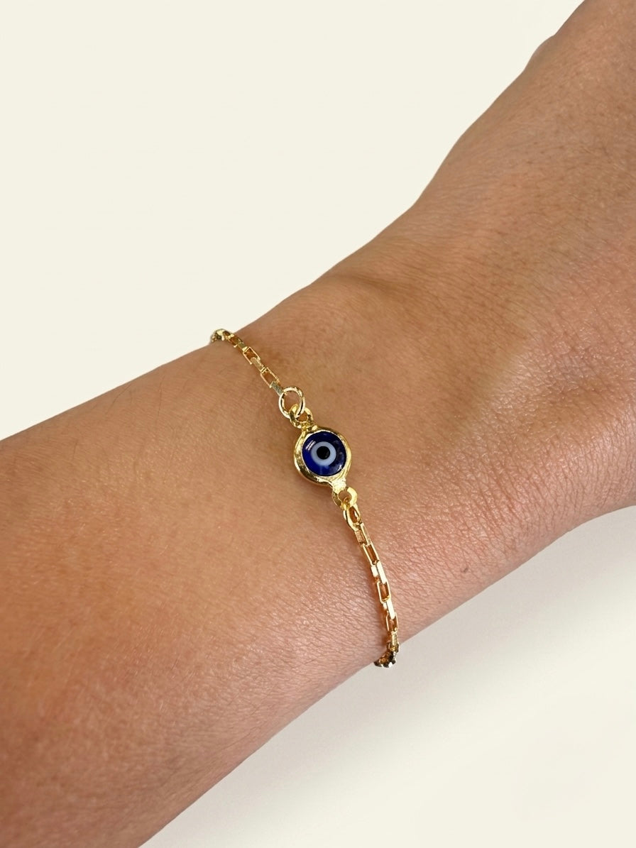 A gold filled paperclip chain bracelet with a blue evil eye charm, shown worn on a person's wrist against a light cream background.