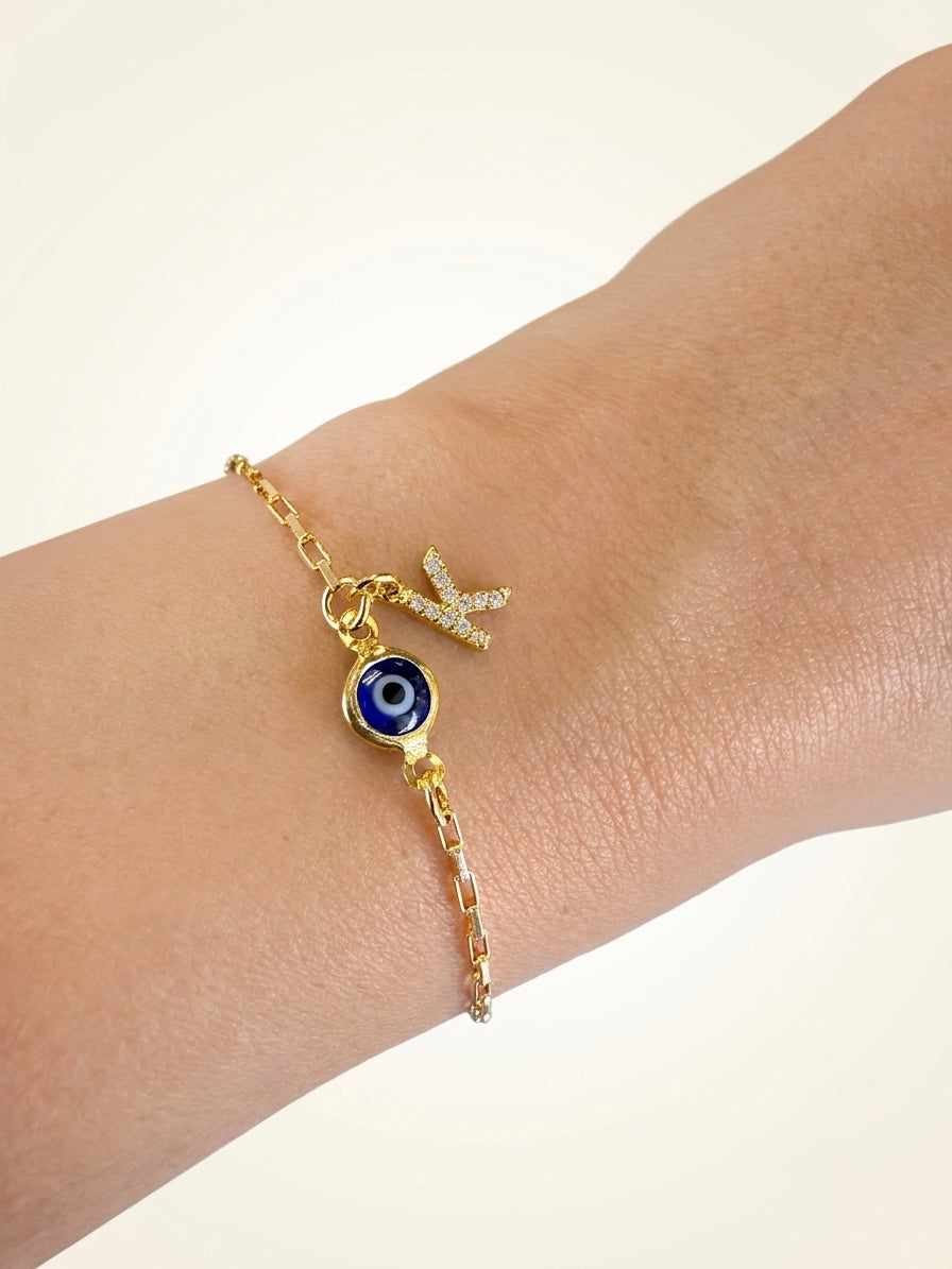 A gold chain bracelet worn on a wrist, featuring a blue evil eye charm and a small gold and cubic zirconia initial charm.
