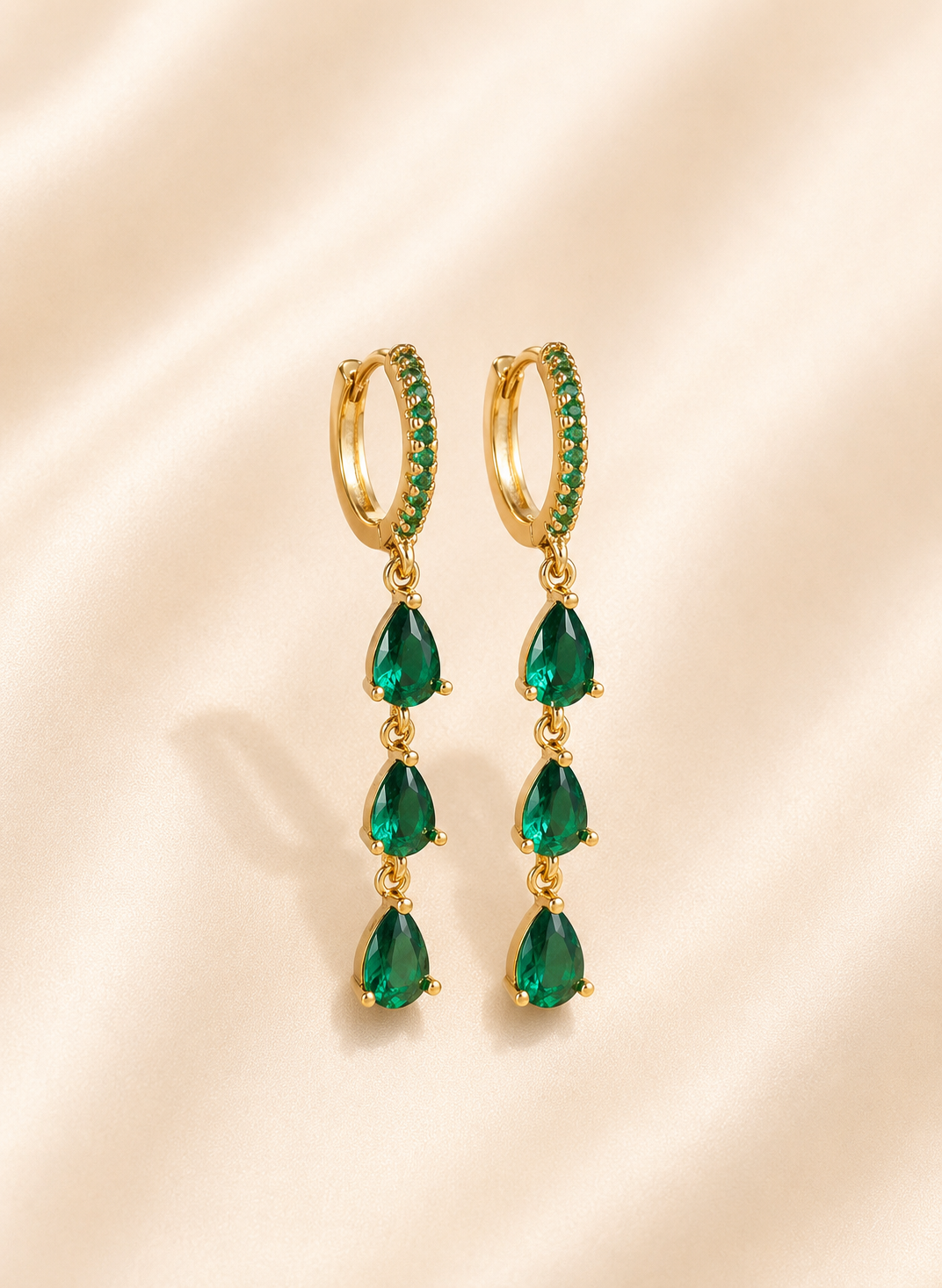 A pair of gold huggie hoop earrings with small green stones along the front, each suspending three teardrop-shaped green gemstones, displayed on a soft beige satin background.