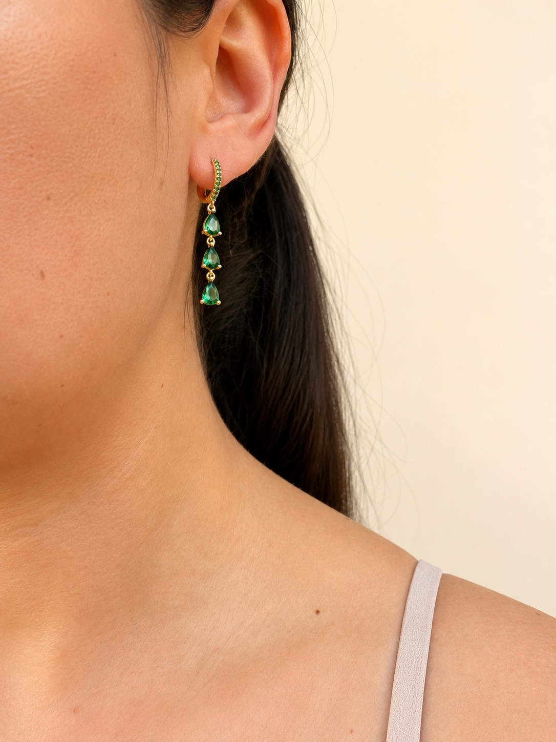 A close-up of a model's ear wearing a gold huggie hoop earring with three dangling green teardrop gemstones.
