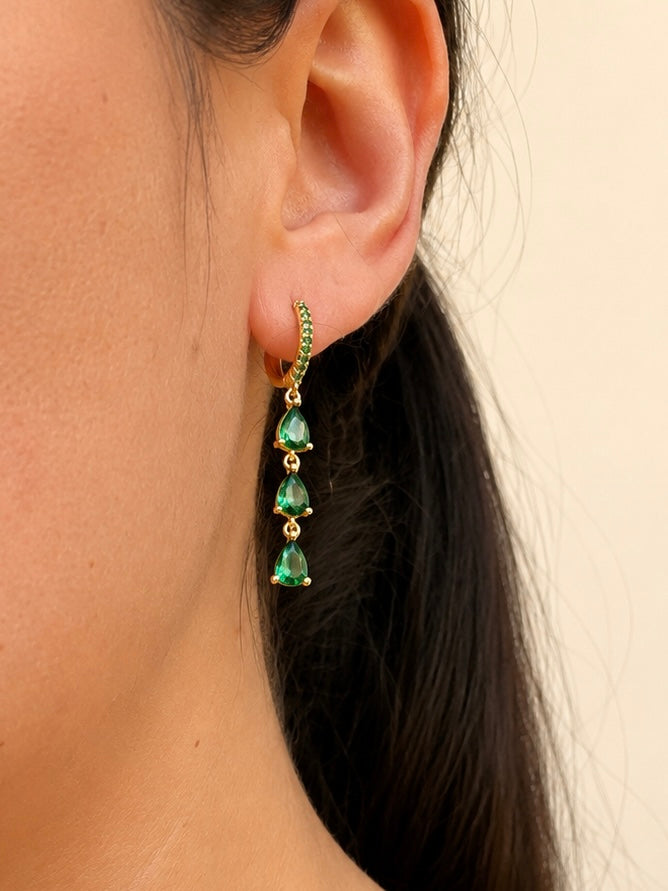 A close-up of a model's ear wearing a gold huggie hoop earring with a vertical drop of four green teardrop gemstones.