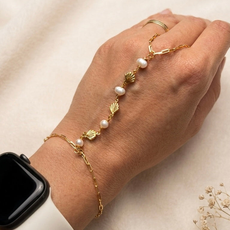 Hand wearing a gold filled hand chain bracelet with pearls and shells on a beige background