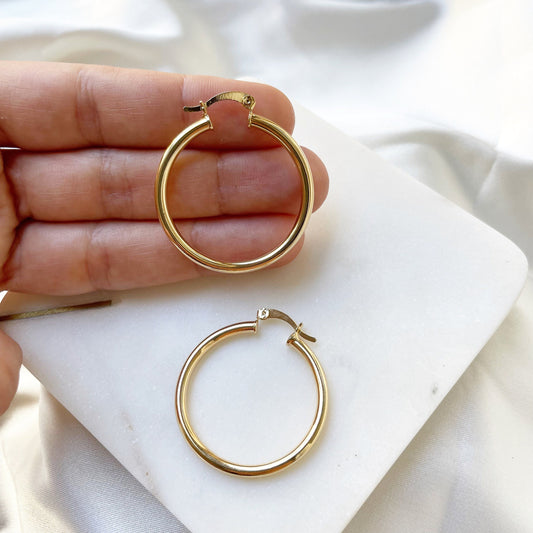 A pair of 18k gold filled thin hoop earrings, displayed on an individual's hand, with a white and gold color scheme.