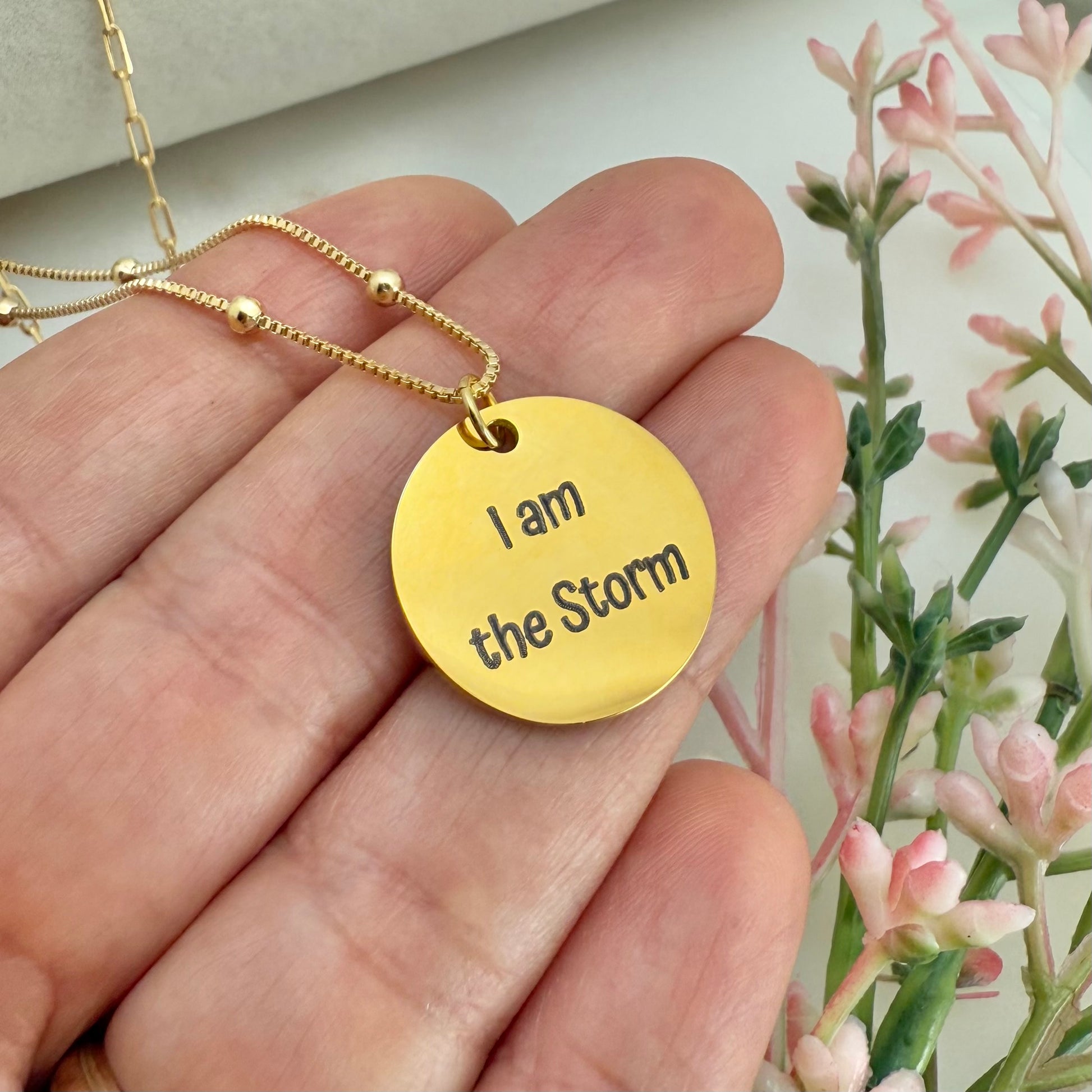 Gold necklace with 'I am the Storm' pendant held in a hand with flowers in the background