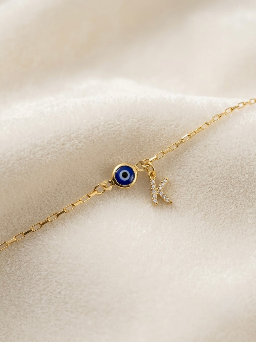 A delicate gold filled chain bracelet featuring a small blue evil eye charm with a small gold initial 'K' charm, displayed on a soft cream-colored fabric background.