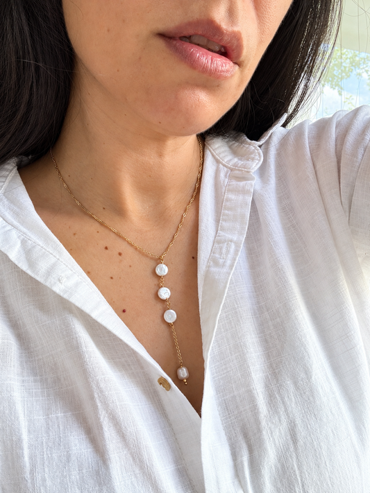 A close-up of a woman wearing a delicate gold lariat necklace with three white pearl-like beads, paired with a white linen button-up shirt.