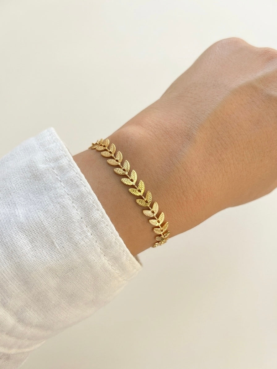 A delicate gold leaf bracelet worn on a person's wrist, shown against a soft cream background with a white linen sleeve visible.