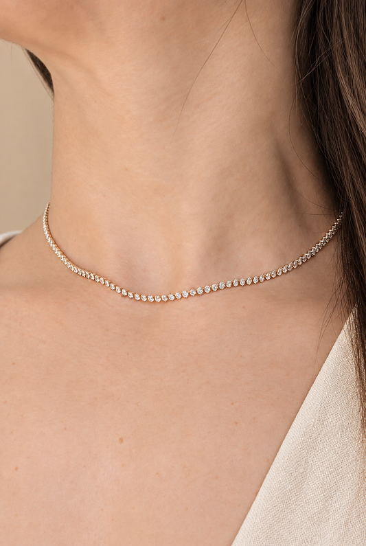 A delicate gold tennis necklace with small clear stones worn on a model's neck.