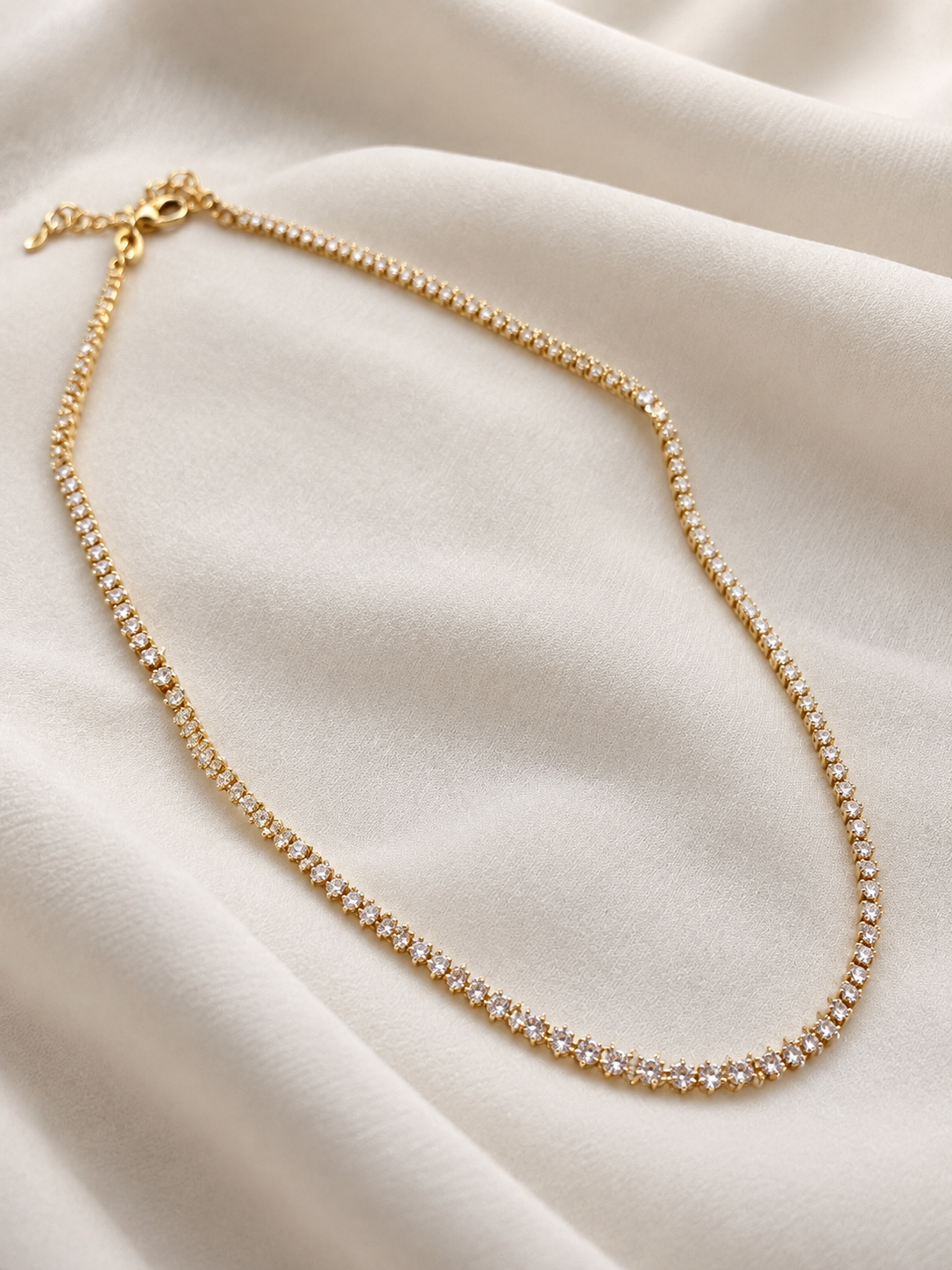 A delicate gold tennis necklace with clear stones, arranged in a U-shape on a draped cream-colored fabric background.
