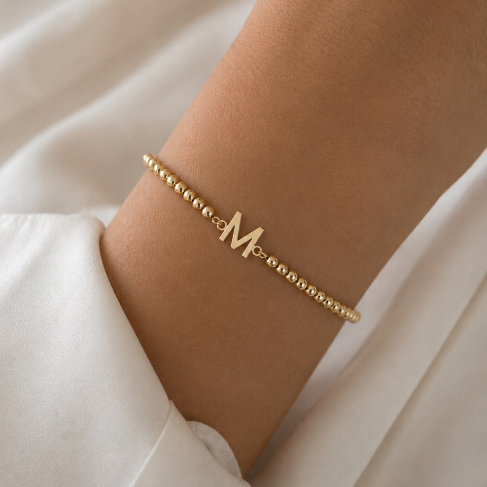 Gold bracelet with letter 'M' on a wrist against a white fabric background