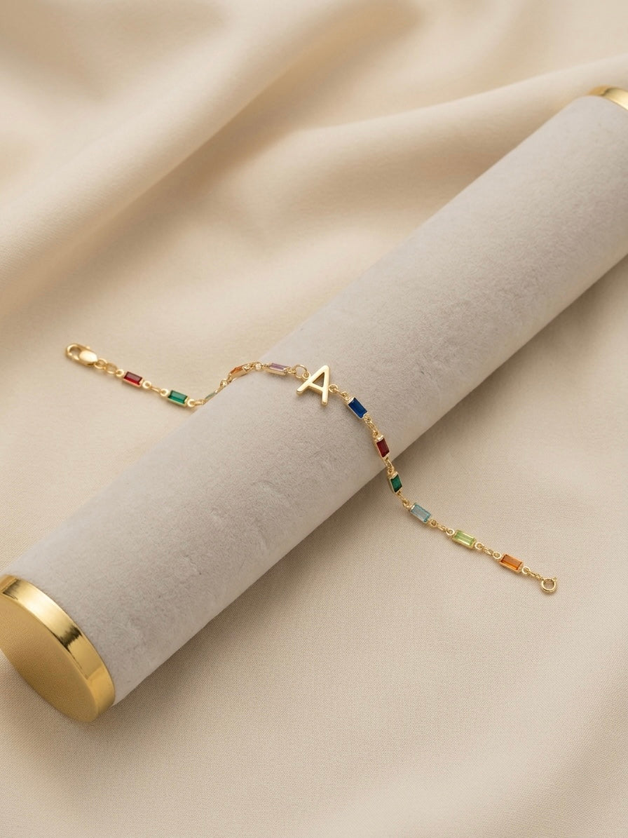 A delicate gold chain bracelet with a small letter 'A' charm and colorful gemstone accents, displayed on a beige velvet jewelry roll against a soft beige fabric background.
