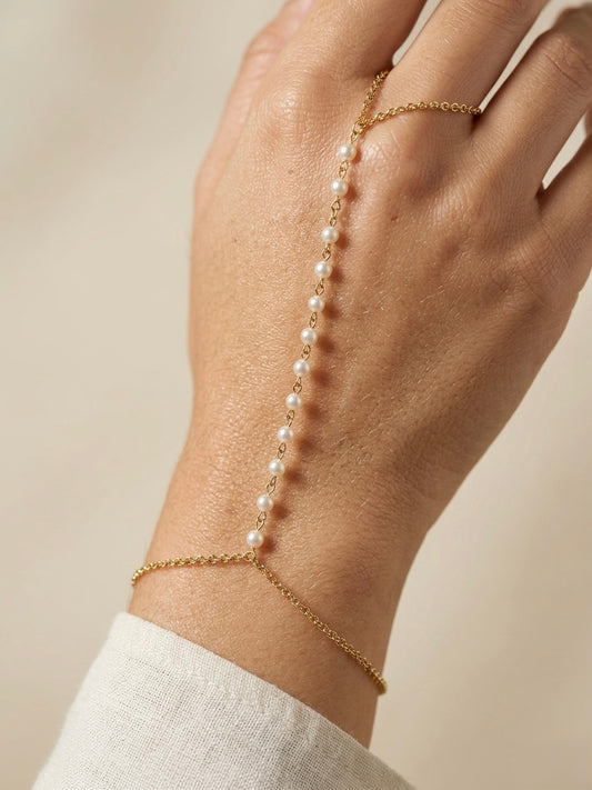 Hand wearing a delicate pearl and gold filled hand chain bracelet on a neutral background