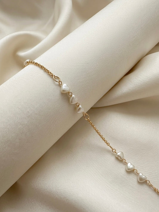 A delicate gold chain bracelet featuring a series of small,  heart shaped white pearl-like beads, elegantly draped over cream-colored satin fabric.