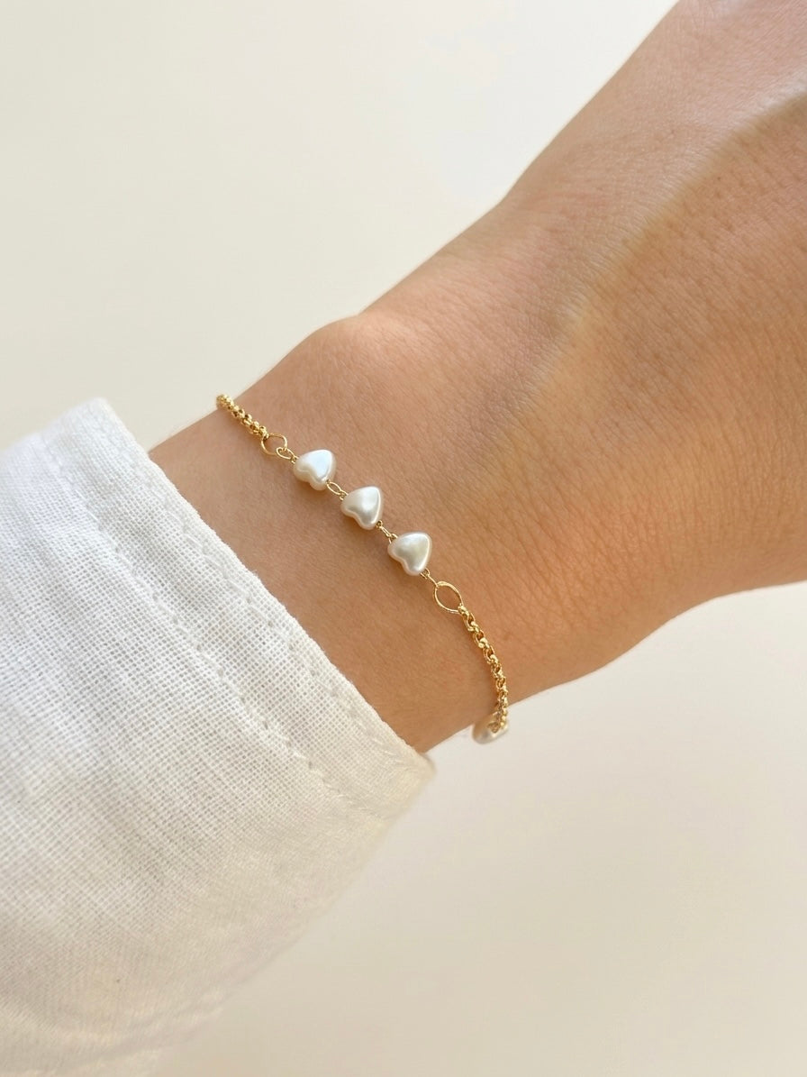 A delicate gold chain bracelet with three small heart shaped white pearl-like beads, shown worn on a person's wrist with a white textured sleeve visible.