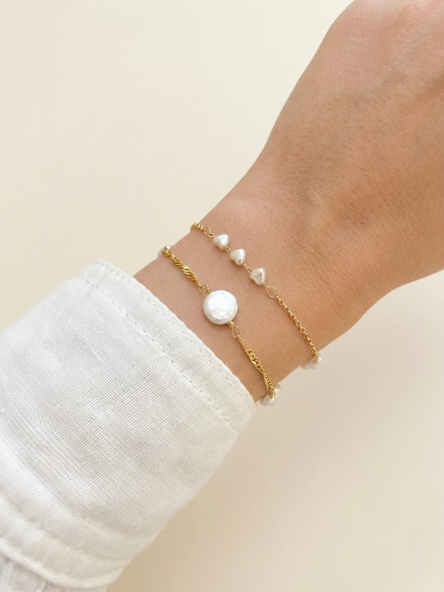 A close-up of a person's wrist wearing three delicate bracelets: two fine gold chain bracelets and one bracelet with small white heart shaped pearls and a layered with a larger round white pearl, against a soft cream background.