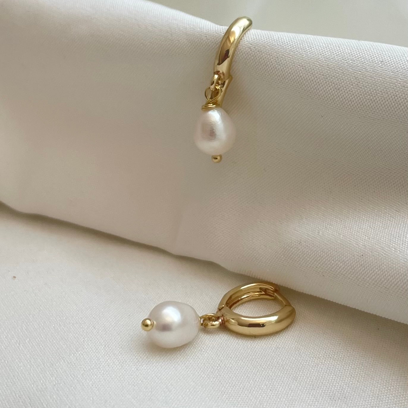 A pair of gold-filled hoop earrings with dangle freshwater pearls, displayed on a white fabric surface.