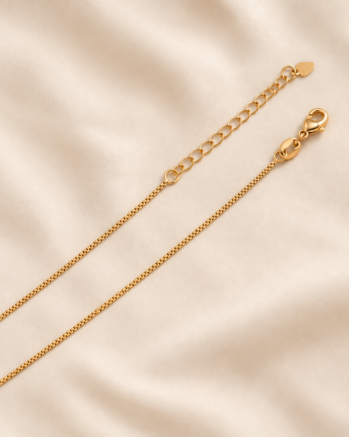 A close-up of a gold chain necklace with an adjustable link extender and lobster clasp, laid diagonally on a soft beige fabric background.