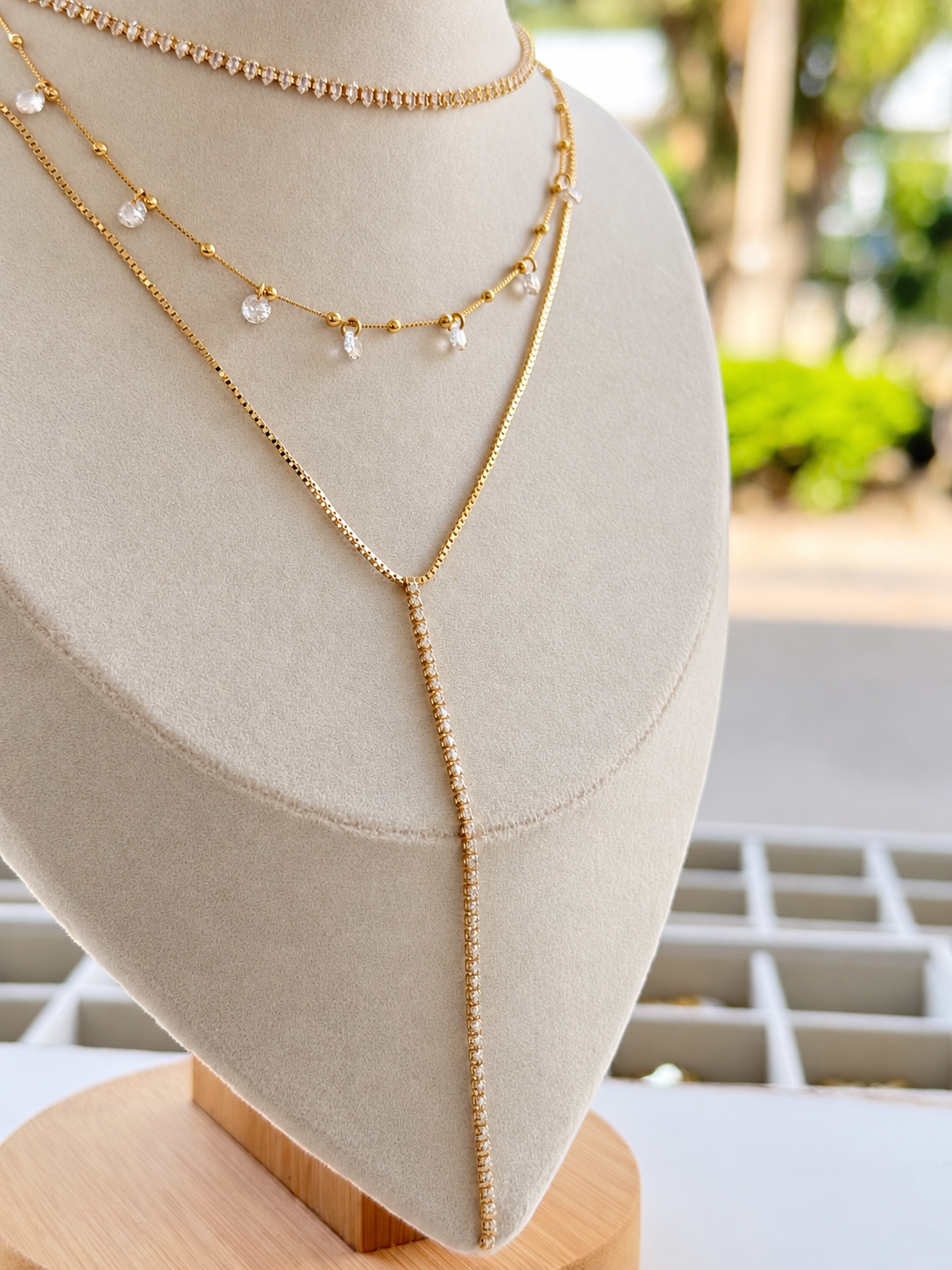 A layered gold necklace set displayed on a beige fabric jewelry bust. The set includes a delicate beaded chain, a chain with small clear crystal charms, and a longer Y-shaped chain draped over the bust.