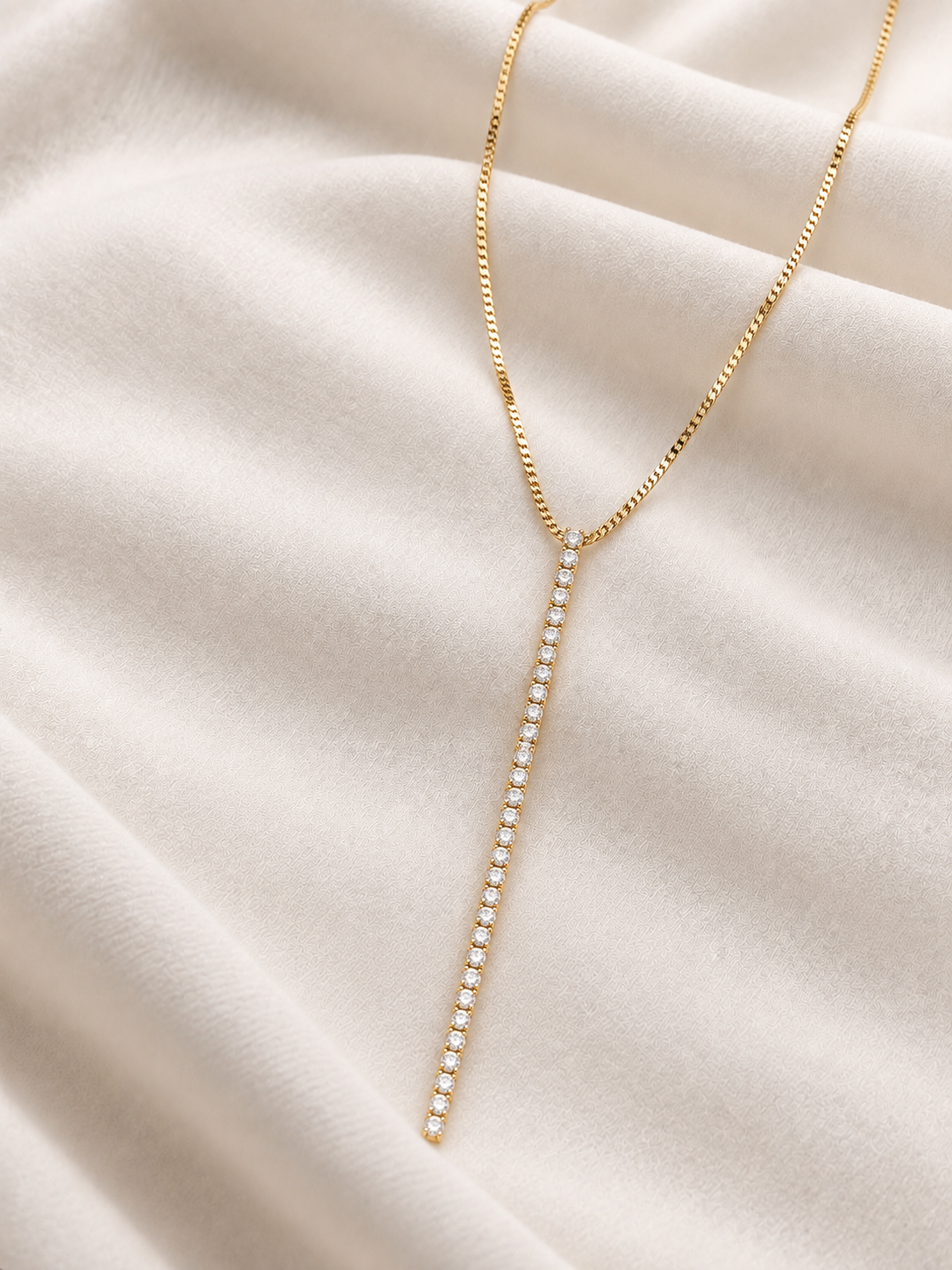 A delicate gold Y-necklace with a vertical drop featuring a line of small clear stones, displayed on draped cream-colored fabric.