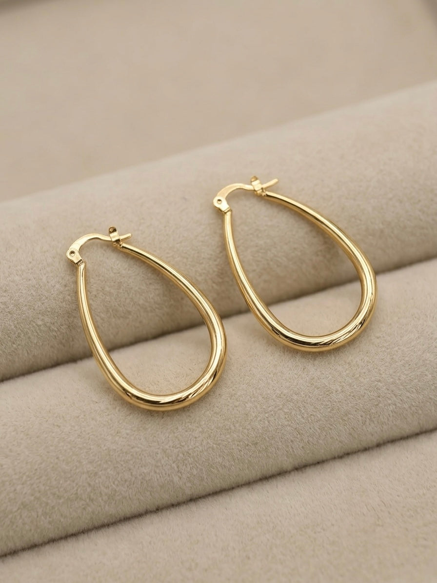 A pair of gold filled teardrop hoop earrings resting on a beige textured jewelry display cushion.