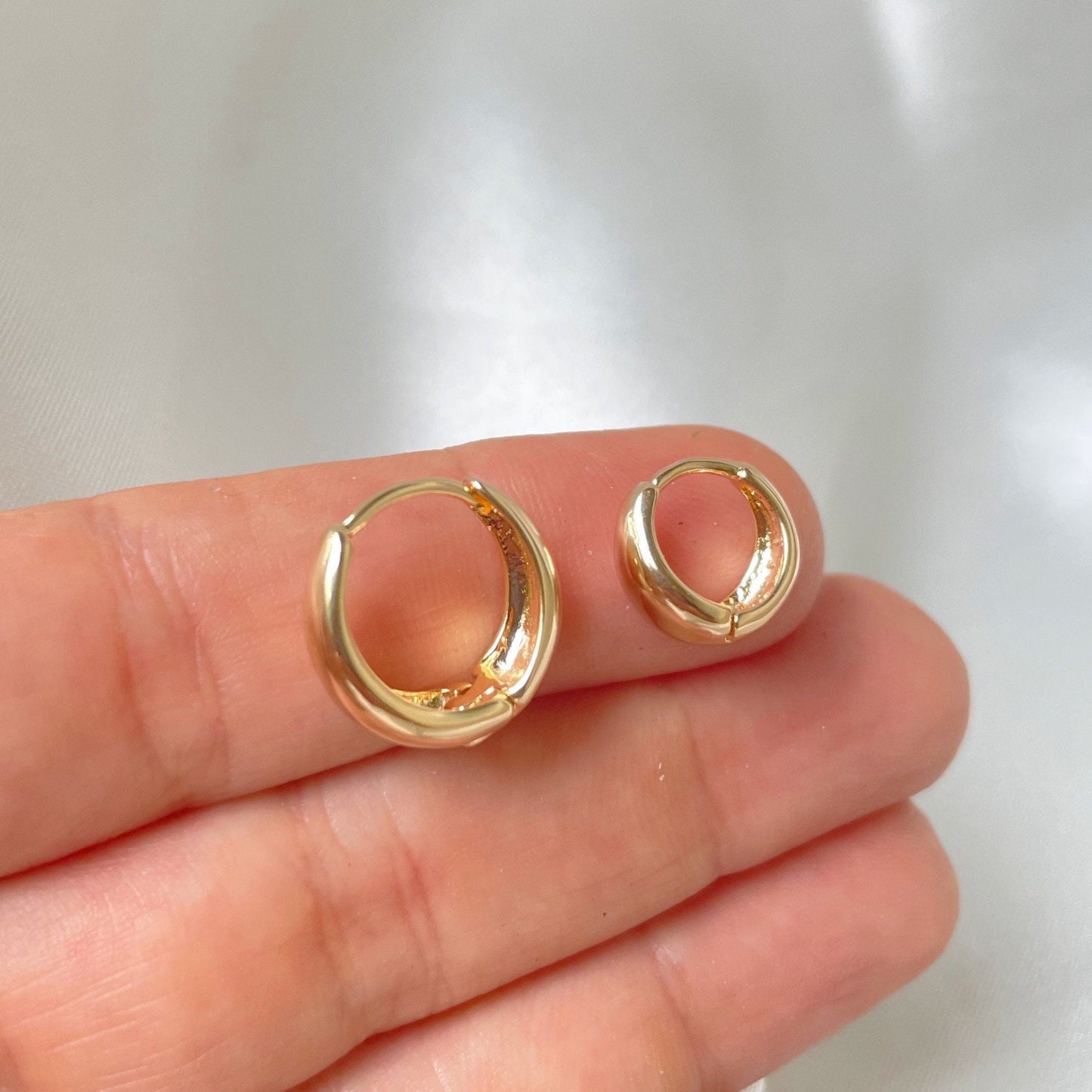A pair of thick gold huggie hoop earrings held between fingers, showcasing their size and design.