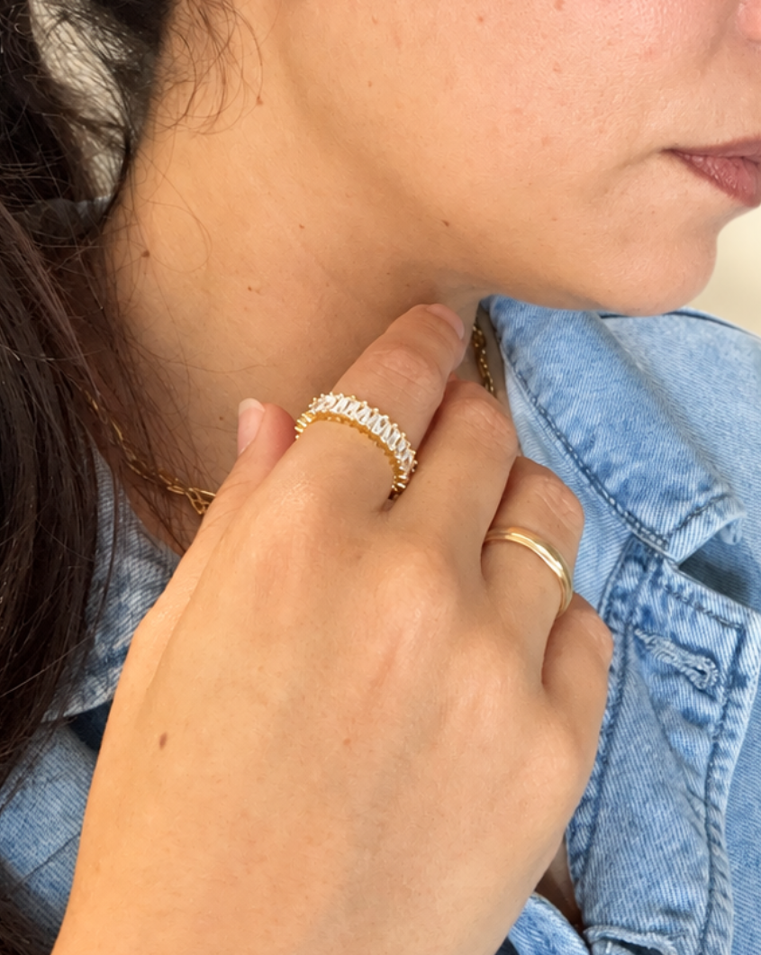 A close-up lifestyle shot of a person wearing a textured gold ring on their index finger and a simple gold band on their middle finger, paired with a denim jacket.