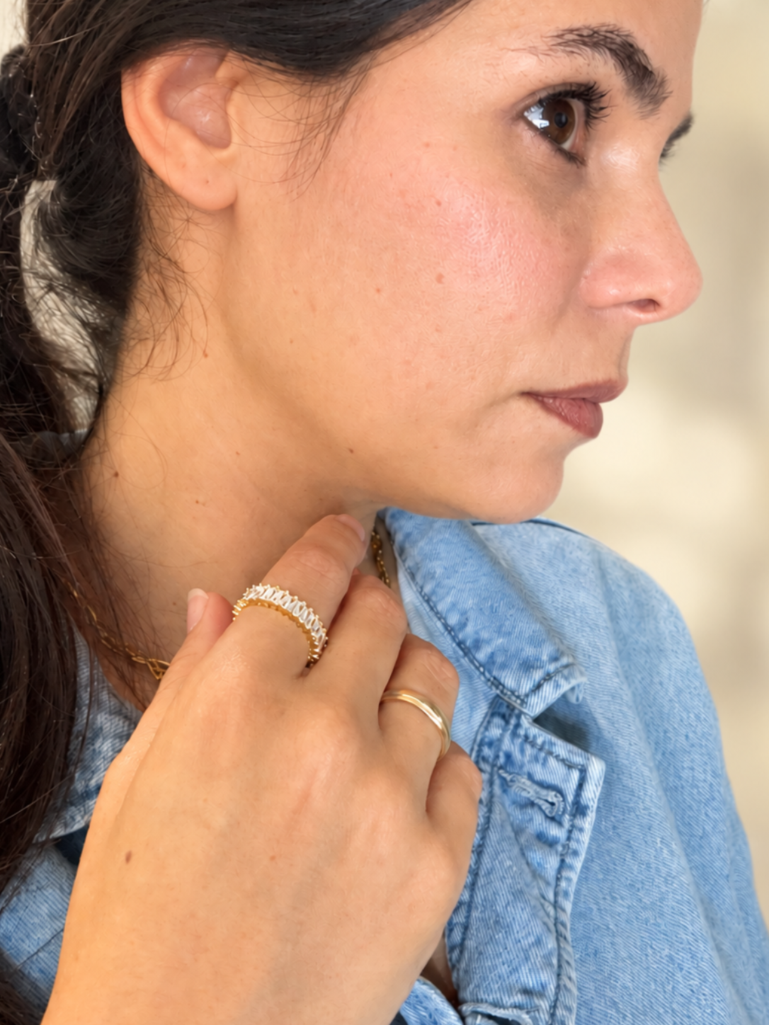 Woman wearing a gold-tone baguette clear crystal ring band with a blue denim shirt against a softly blurred cream background.