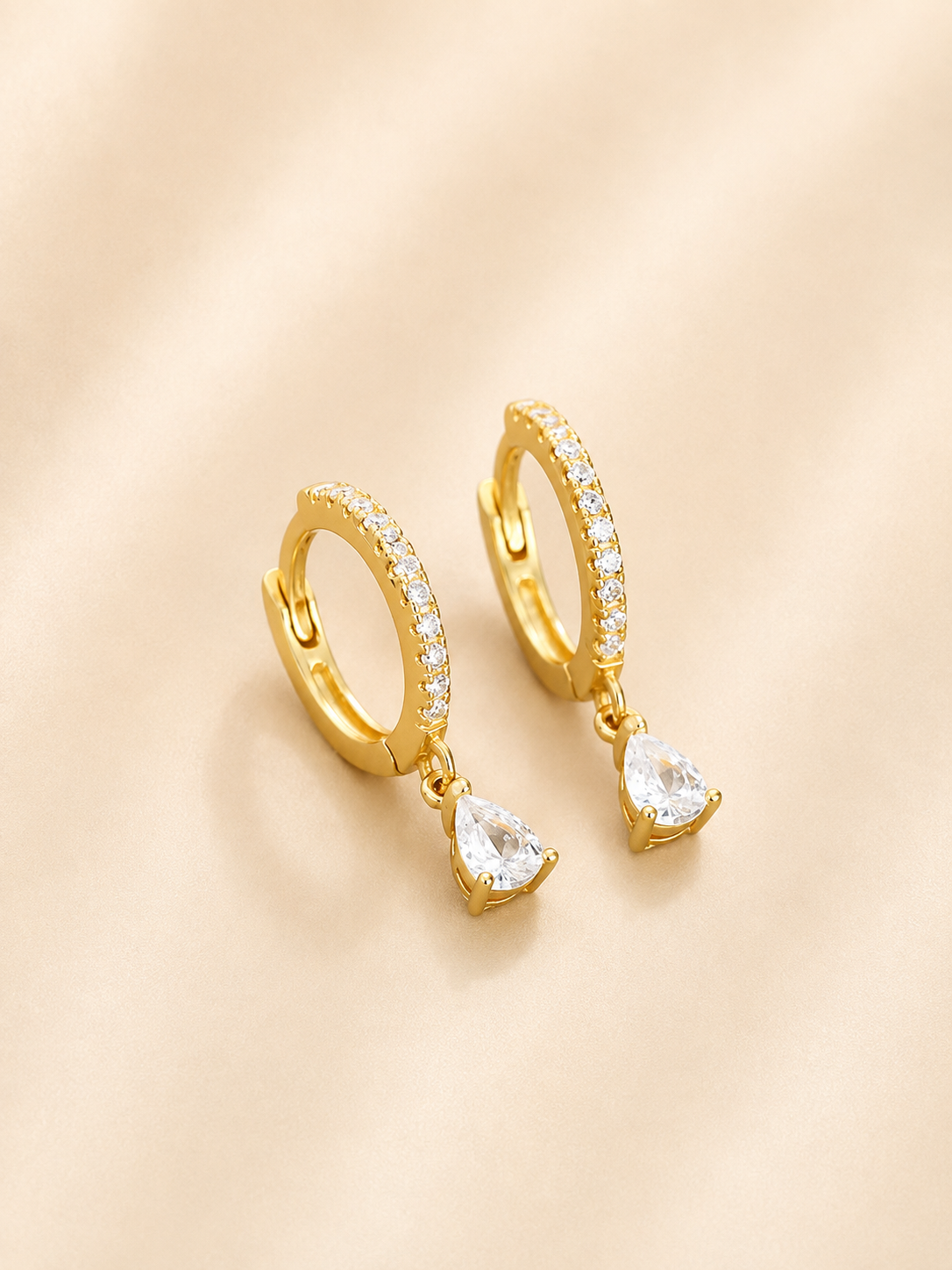 A pair of gold huggie hoop earrings with small clear stones along the top band and a dangling pear-cut clear crystal, displayed on a soft beige fabric background.