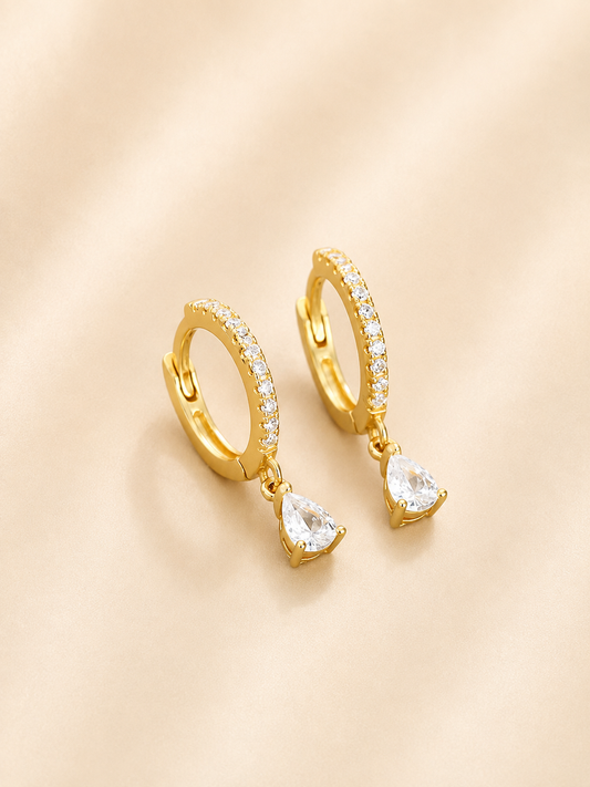 A pair of gold huggie hoop earrings with small clear stones along the top band and a dangling pear-cut clear crystal, displayed on a soft beige fabric background.