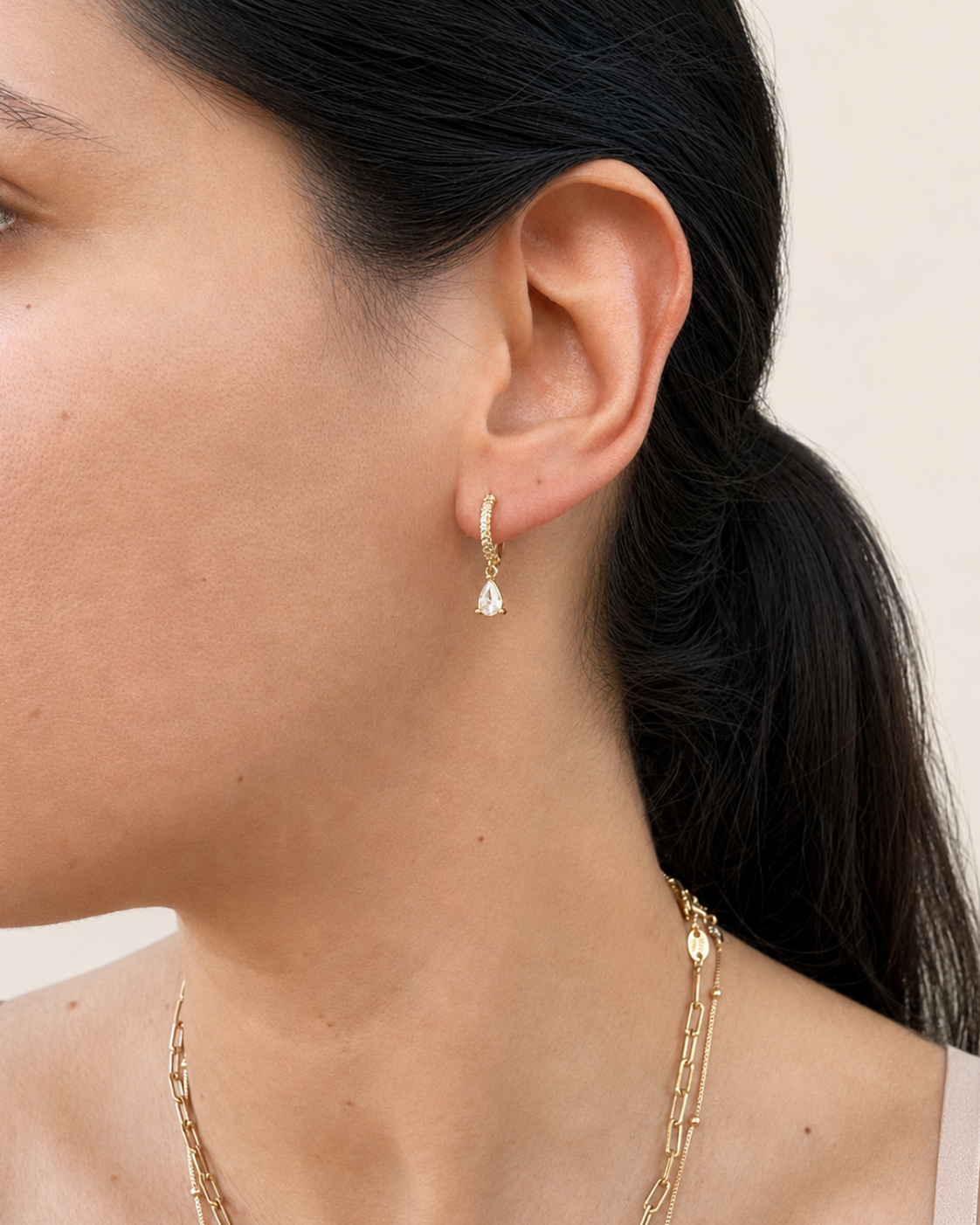 A close-up side profile of a woman wearing a delicate gold huggie hoop earring with a small teardrop crystal drop, paired with layered gold necklaces against a soft beige background.