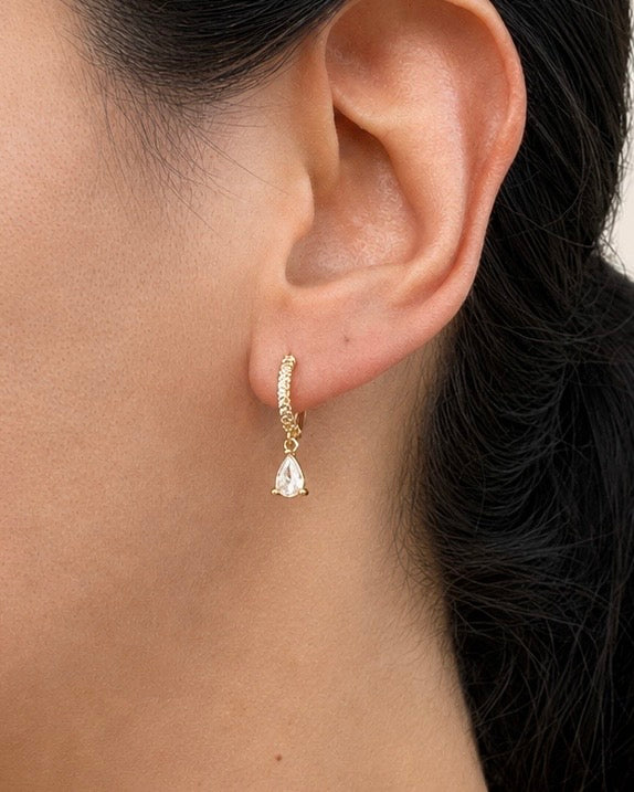 A close-up of a model's ear wearing a delicate gold huggie hoop earring with a small clear teardrop gemstone dangle.