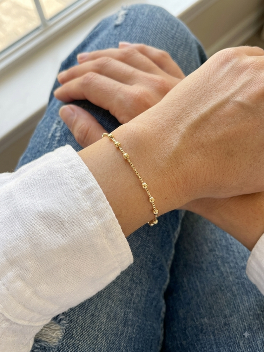 A delicate gold beaded chain bracelet worn on a person's wrist, shown in a close-up lifestyle shot with blue jeans and a white shirt visible.
