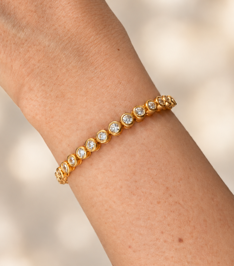 close up of A gold tennis bracelet with small clear gemstones worn on a person's wrist, shown against a softly blurred light beige background.